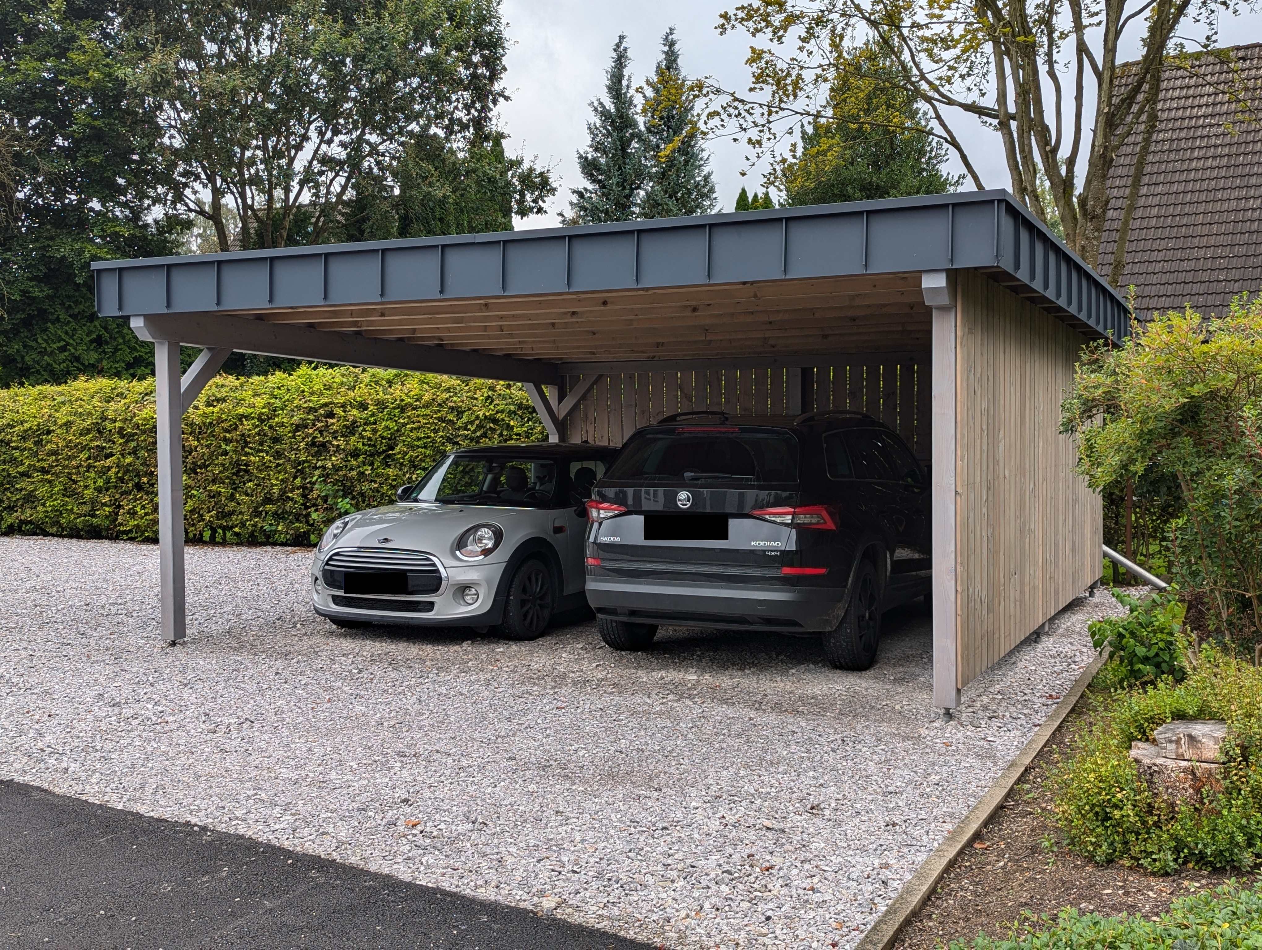 Holzcarport mit zwei parkenden Fahrzeugen auf Kiesfläche vor Hecke und Wohnhaus